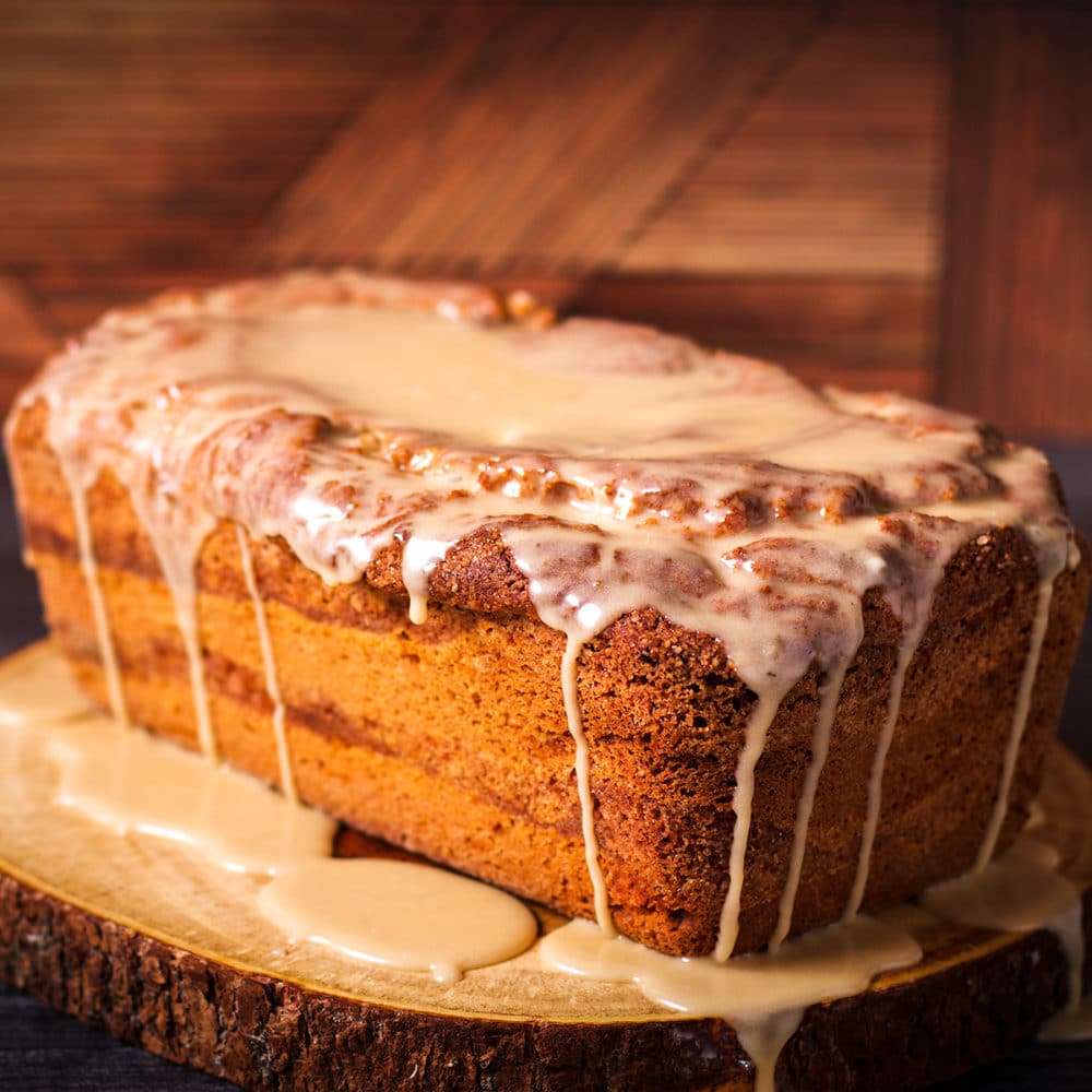 A cinnamon loaf cake covered in vanilla icing sitting on a wood cutting board.