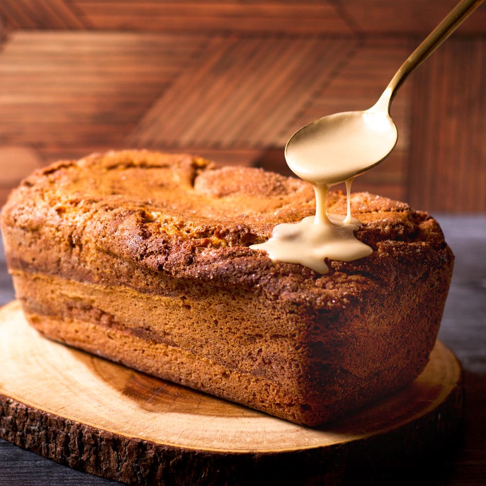 Someone using a gold spoon to drizzle vanilla icing over the top of a cinnamon loaf cake.