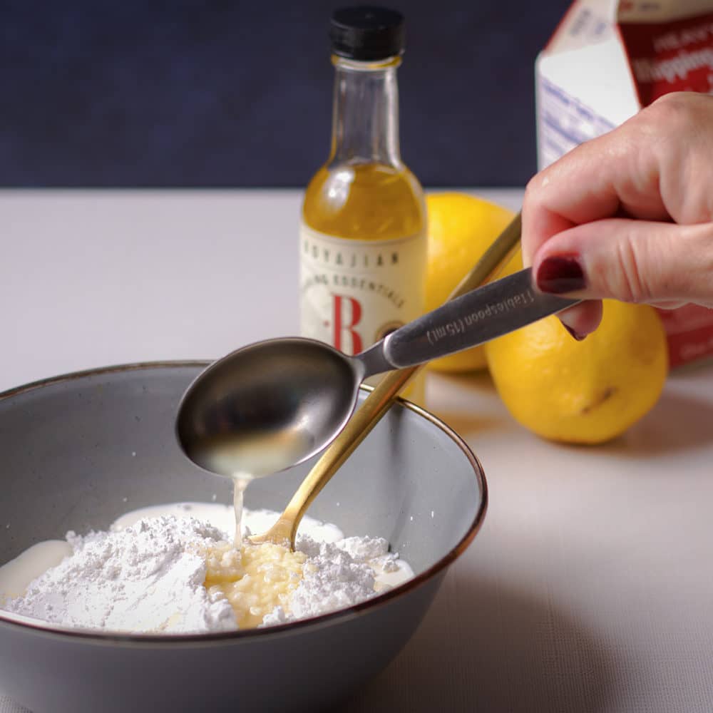 Someone pouring a tablespoon of lemon juice into a bowl of powdered sugar.
