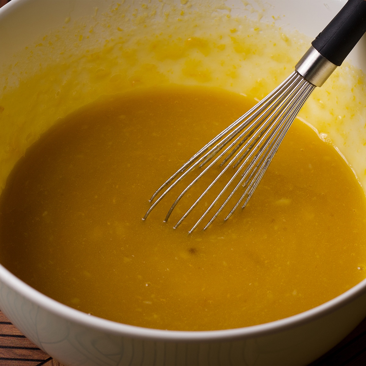 Someone using a wire whisk to mix melted butter, olive oil, honey, whole eggs and an egg yolk, lemon zest, almond extract, and sour cream in a white bowl.