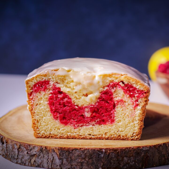 A raspberry swirl loaf cake on a wood cutting board that's been cut in half so you can see the bright red ribbon of raspberry cake swirled into the loaf.