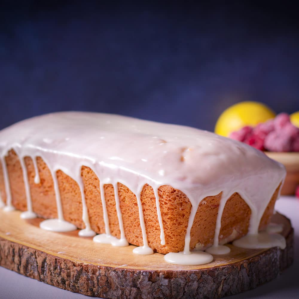 A raspberry loaf cake covered in vanilla icing on a wood cutting board with fresh lemons and raspberries in the background.