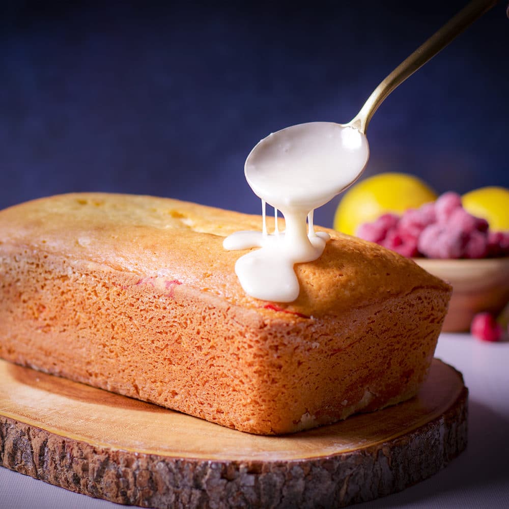 Someone using a gold spoon to top a raspberry loaf cake with lemon icing.
