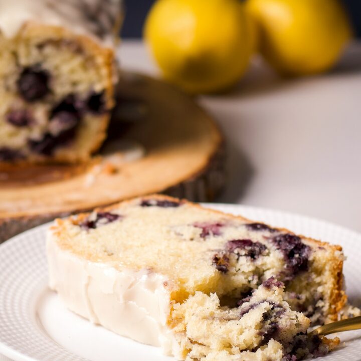 A slice of blueberry loaf cake with lemon icing on a white plate with a gold fork taking a bite of the cake.