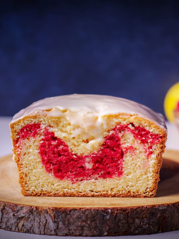 A raspberry swirl loaf cake on a wood cutting board that's been cut in half so you can see the bright red ribbon of raspberry cake swirled into the loaf.