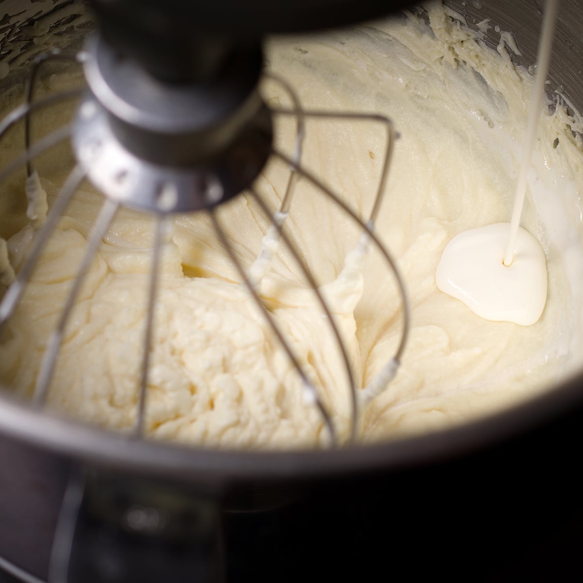 Someone pouring cream into the bowl of an electric mixer that's beating white chocolate ganache.
