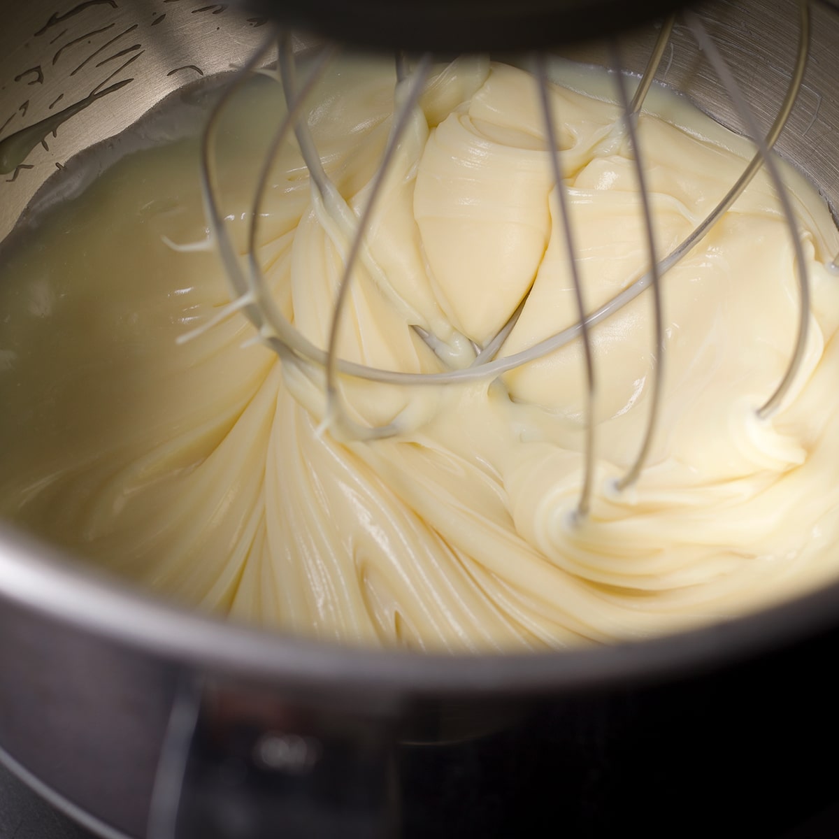 Looking down into the bowl of an electric stand mixer while using the whisk attachment to beat white chocolate ganache.
