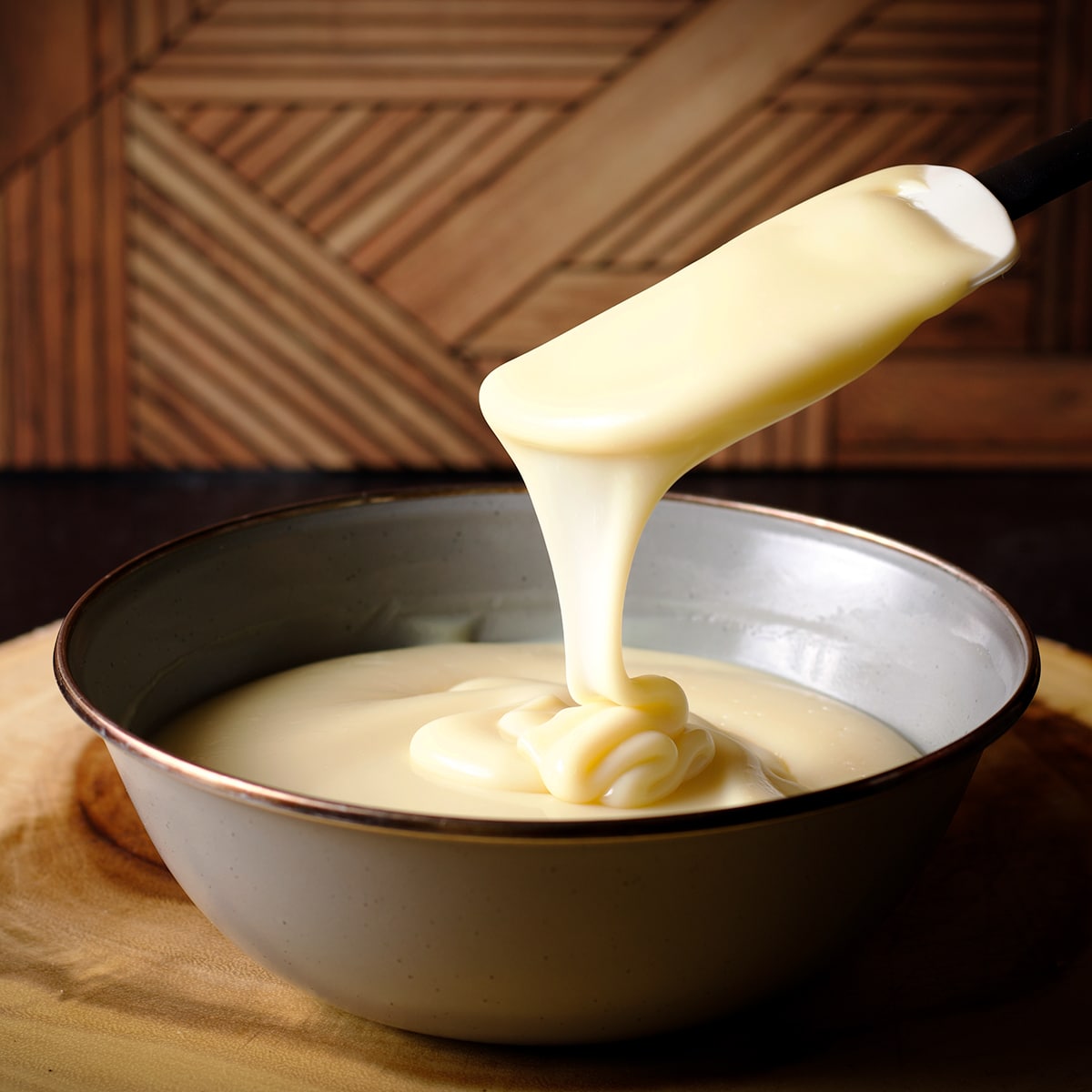 Someone lifting a rubber spatula from a bowl of white chocolate ganache so the ganache is dripping from the spatula back into the bowl in smooth, creamy ribbons.