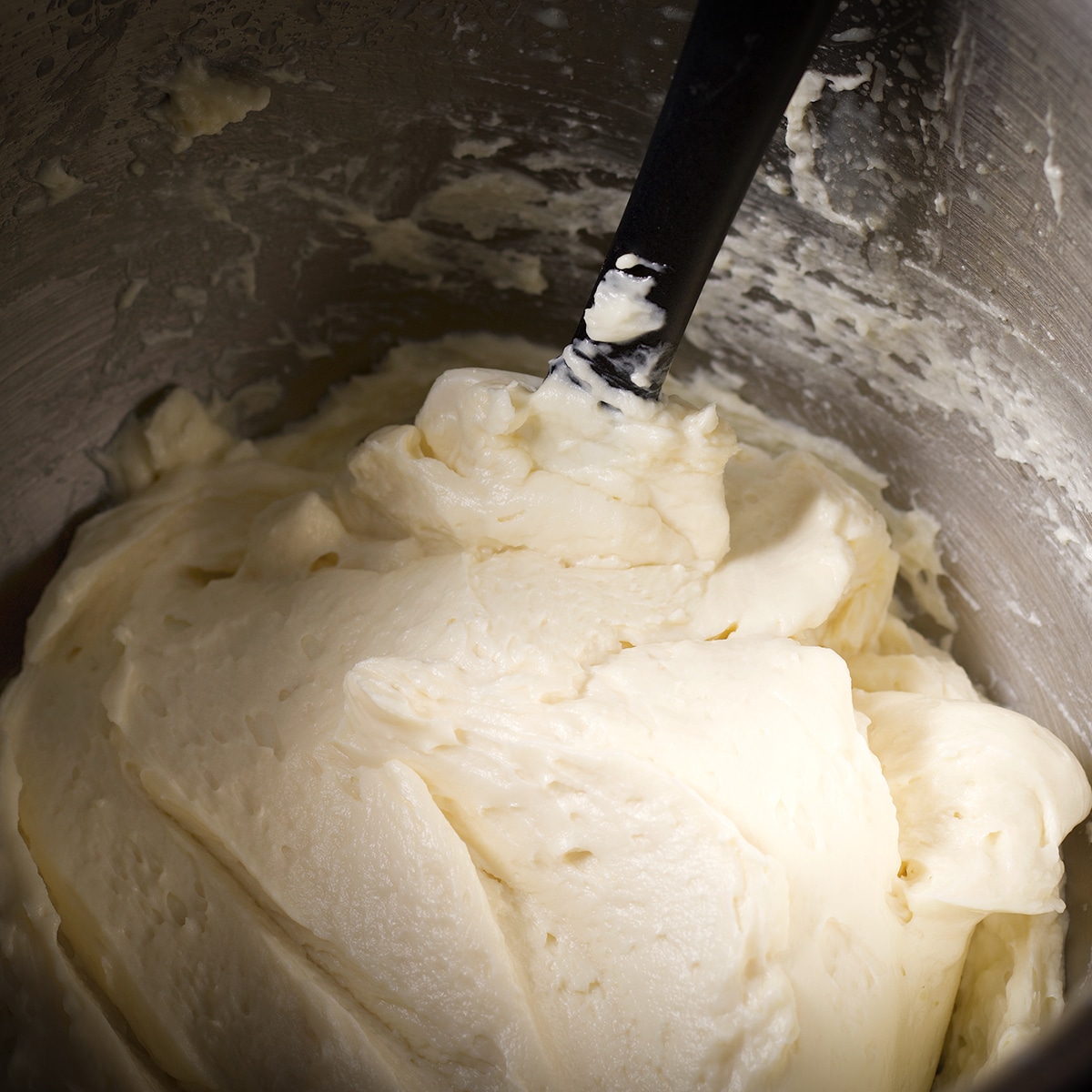 A mixing bowl filled with white chocolate ganache buttercream. A spatula is in the buttercream and you can see how smooth and creamy the frosting is.