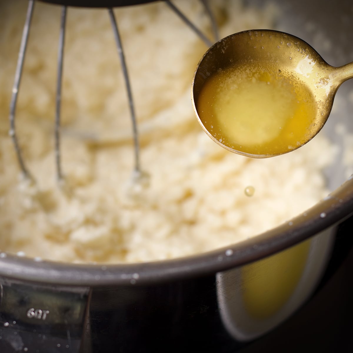 Someone using a gold spoon to slowly add melted butter to white chocolate ganache buttercream while an electric mixer beats the buttercream.