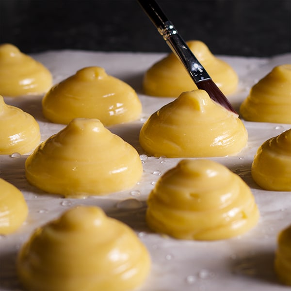 Someone using a pastry brush to coat the tops of profiteroles with an egg wash before baking.