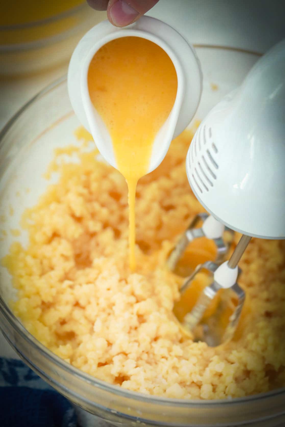 Pouring beaten eggs into choux pastry dough while using an electric mixer to beat the dough.