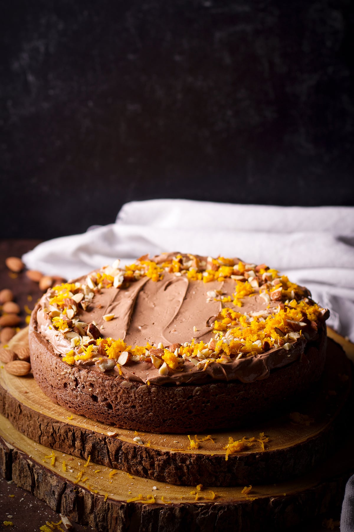 A one layer chocolate almond cake on a wood cake tray. The cake has been frosted with chocolate orange buttercream and decorated with chopped almonds and orange zest.