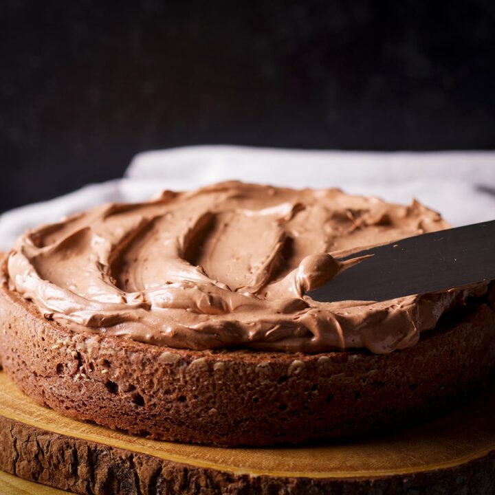 Someone using an icing spatula to spread chocolate orange buttercream over a chocolate cake.
