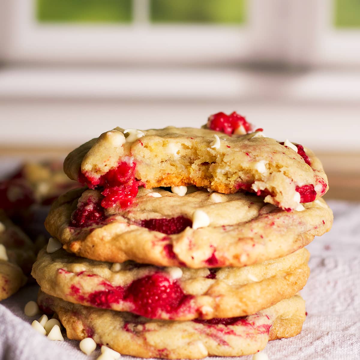 A close up photo of a stack of four white chocolate raspberry cookies. The top cookie has a bite taken out of it so you can see the chewy middle of the cookie.