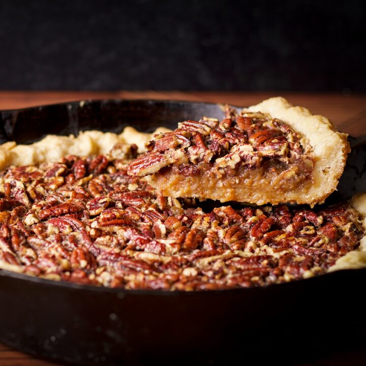 Someone using a pie server to lift a slice of pecan pie from a cast iron skillet.