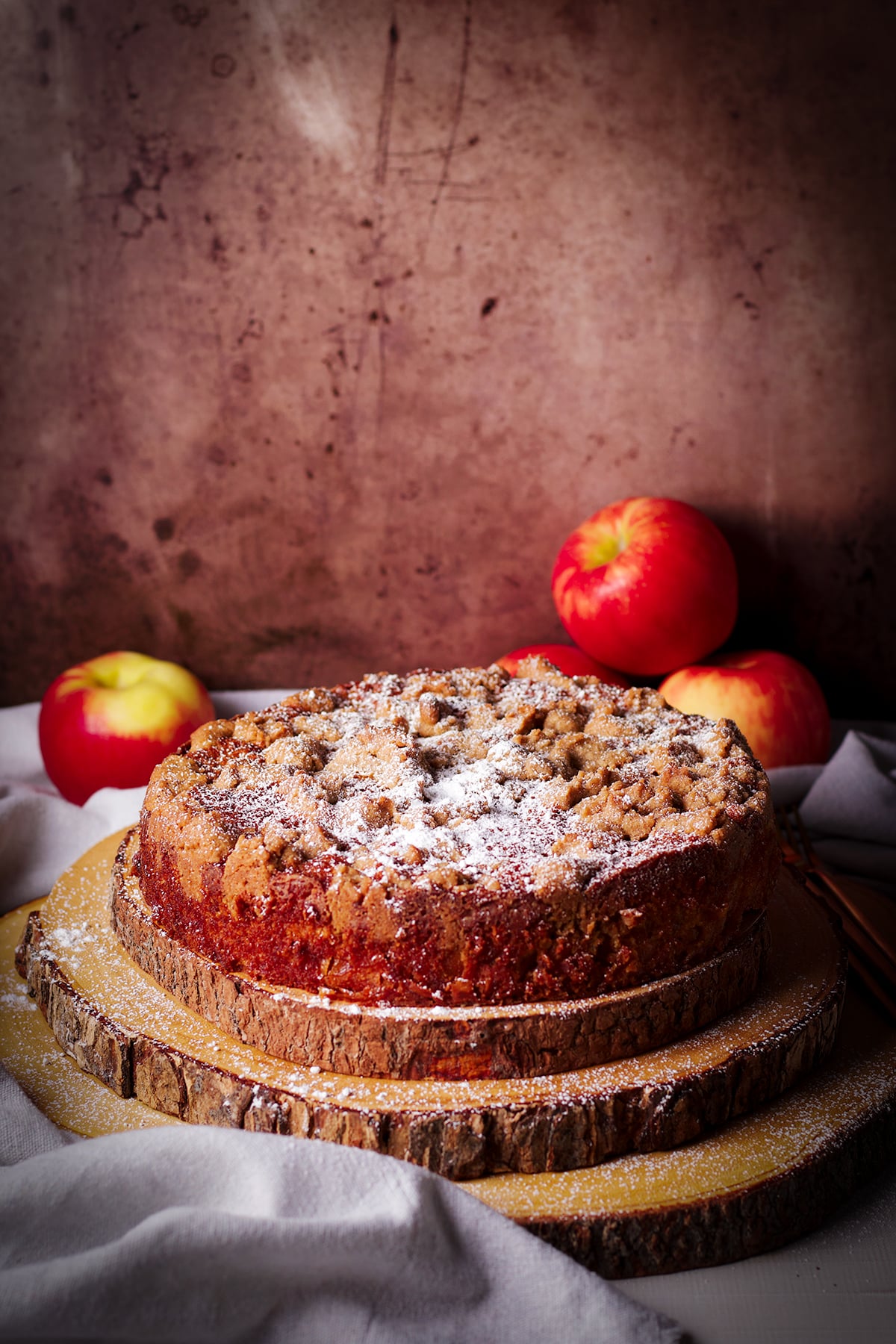 A French Apple Crumb Cake dusted with powdered sugar on a wood platter with red apples piled on the table next to it.