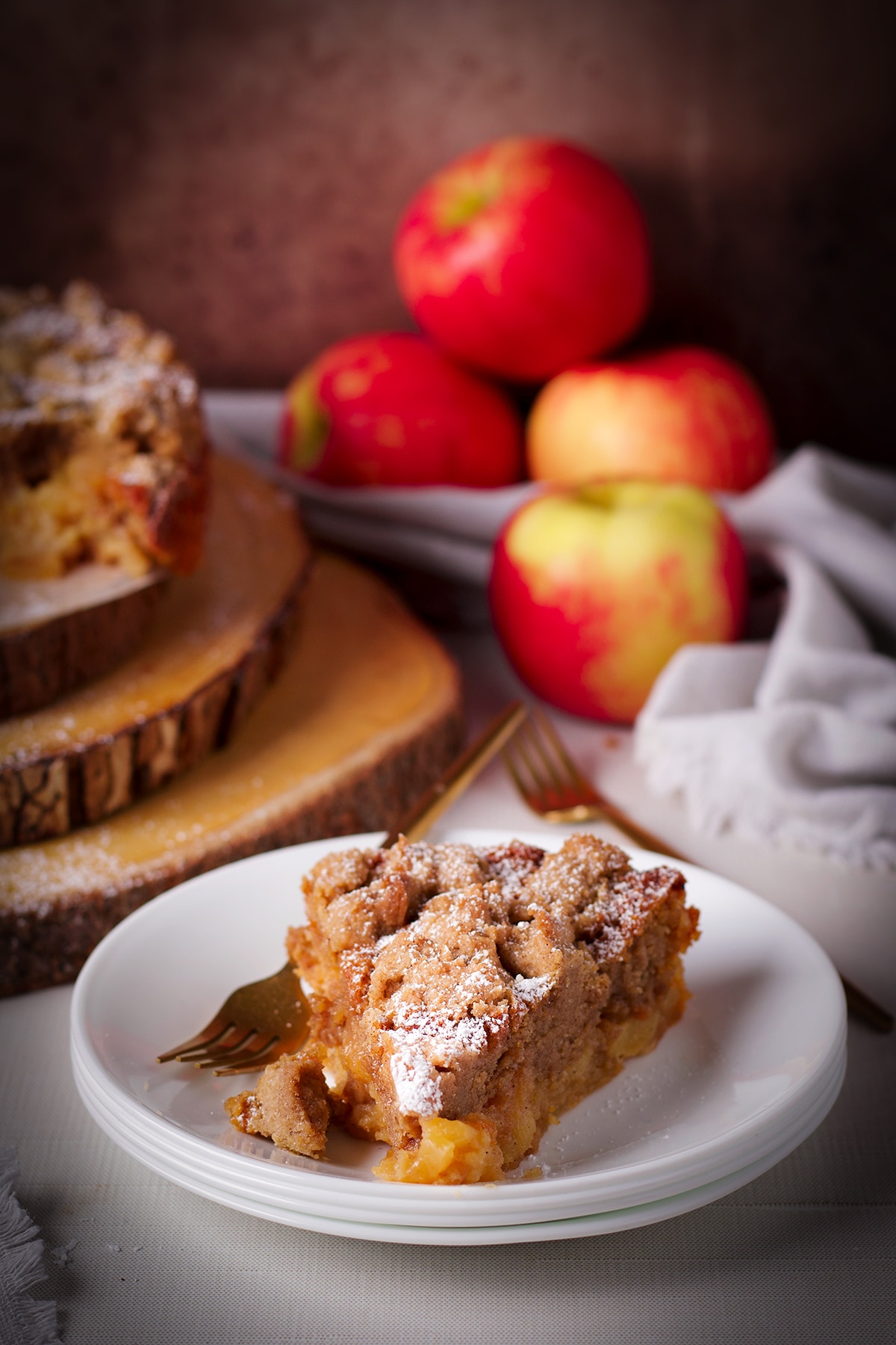 A slice of French Apple Crumb Cake with apples and the rest of the cake in the background.