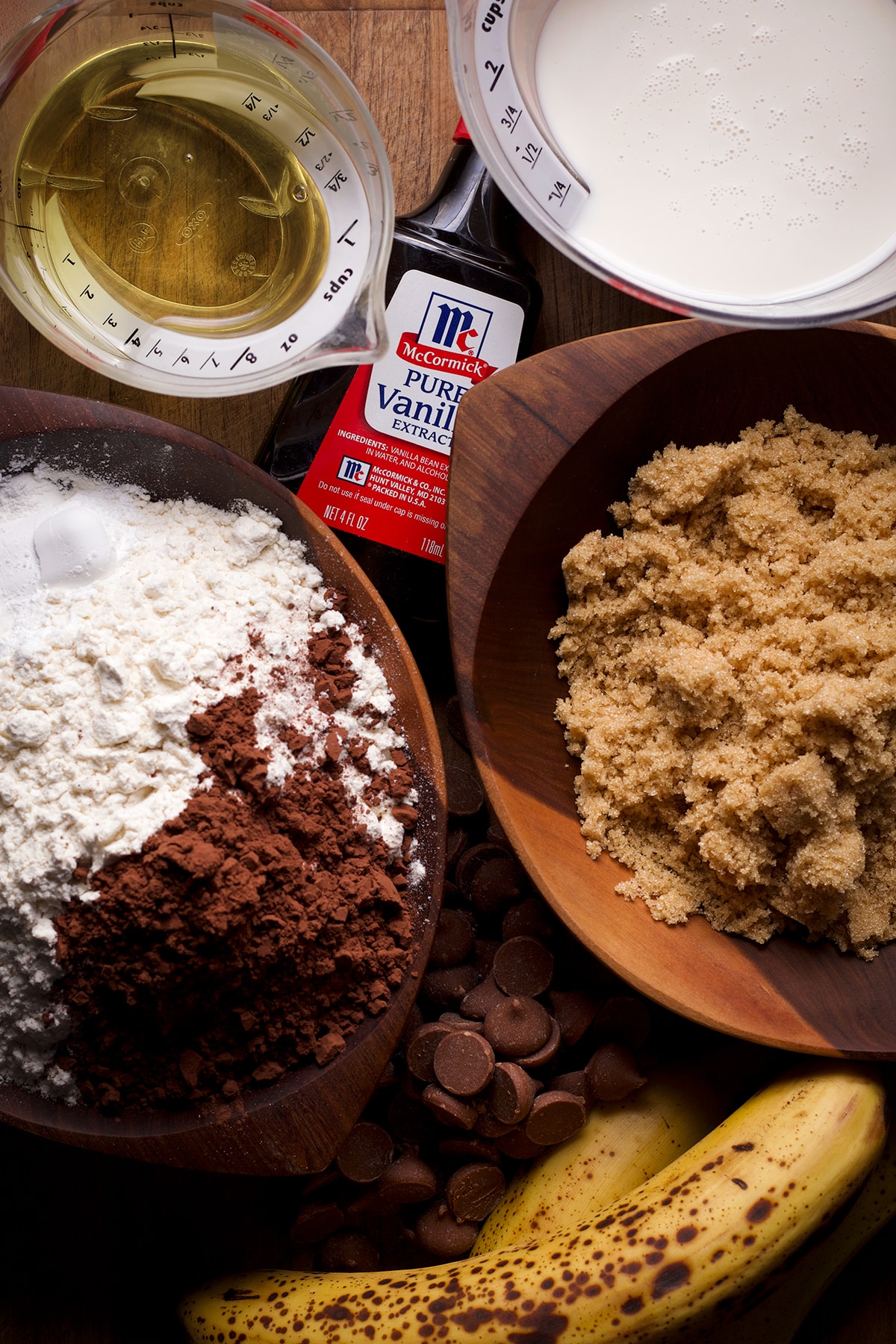 All the ingredients needed to make vegan chocolate banana muffins set out on a wood cutting board.