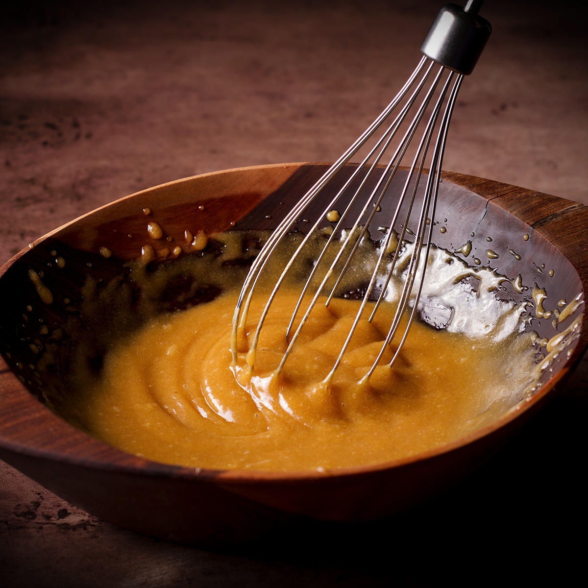 Using a wire whisk to mix the ingredients for maple tahini glaze in a wooden bowl.