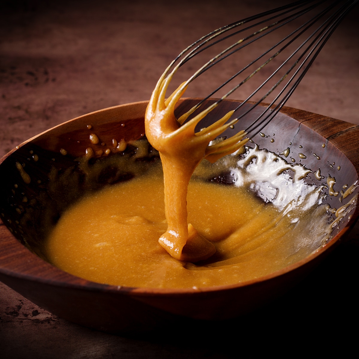 Someone lifting a wire whisk from a bowl of maple tahini glaze and the glaze is falling from the whisk into the bowl like a thick ribbon.