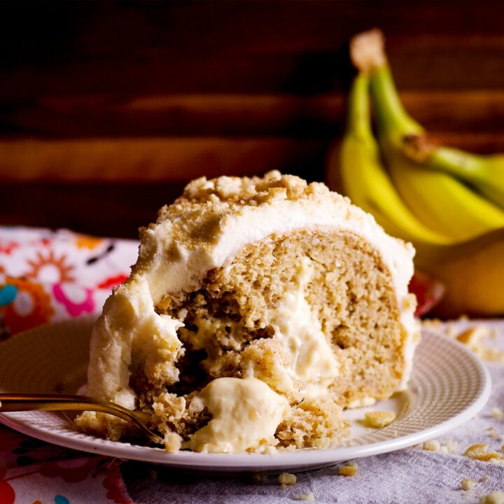 A slice of banana cream cake on a white plate with a bunch of bananas in the background. A gold fork is cutting a bite from the slice of cake.