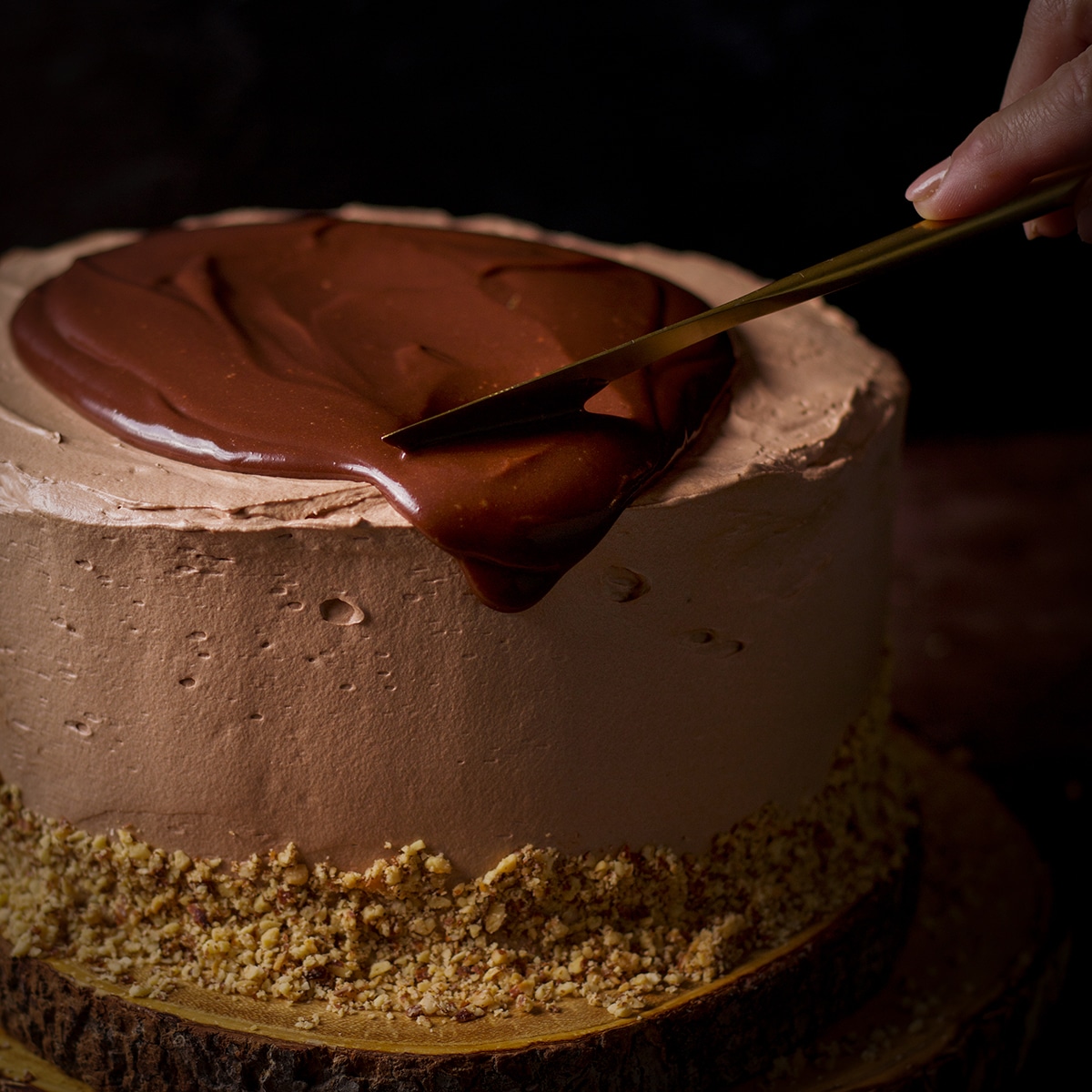 Someone using a knife to spread dark chocolate ganache over the top of a chocolate truffle cake.