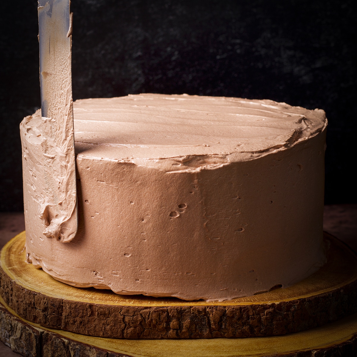 Someone using an icing spatula to smooth milk chocolate buttercream over the sides of a 2-layer chocolate truffle cake.
