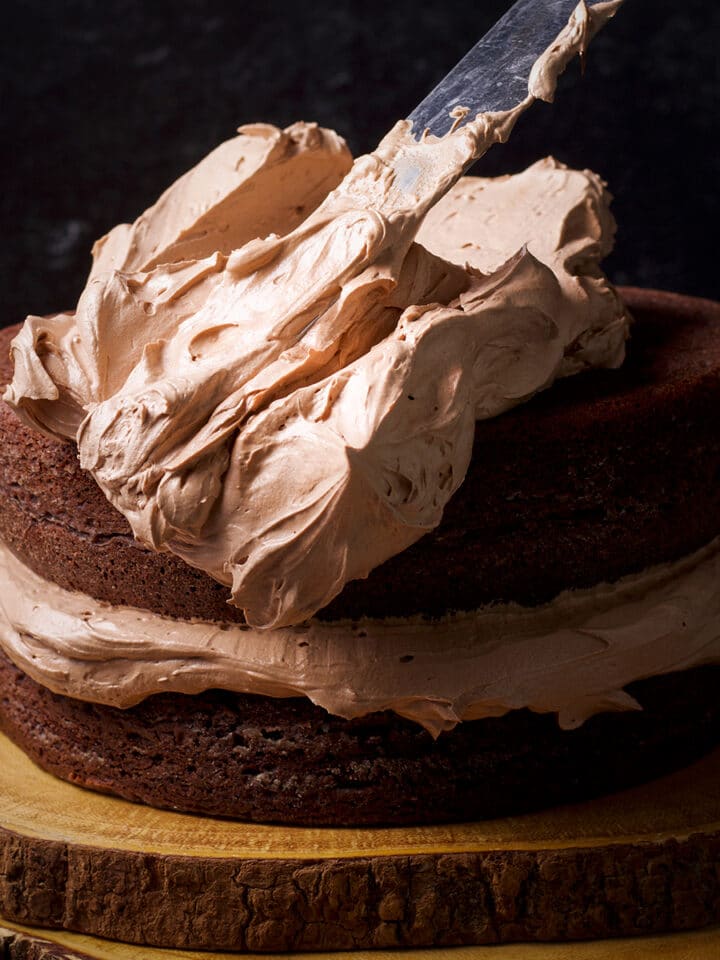 Someone using an offset icing spatula to spread milk chocolate buttercream over the top of a two-layer dark chocolate cake.