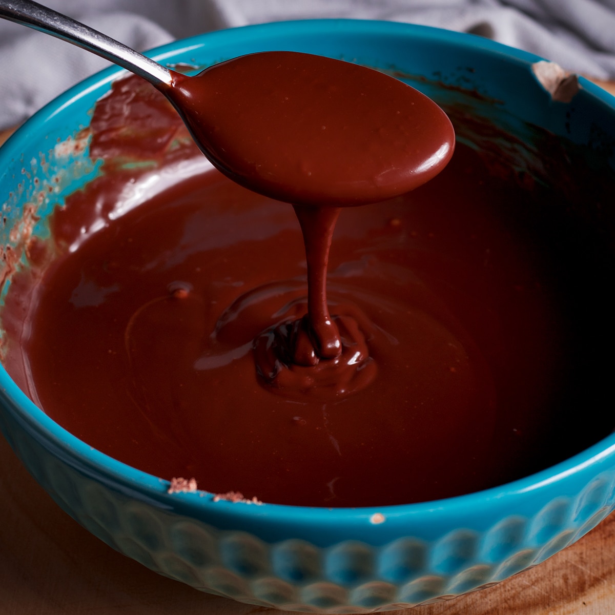 Someone lifting a spoon from a bowl of chocolate ganache so you can see how smooth it is.