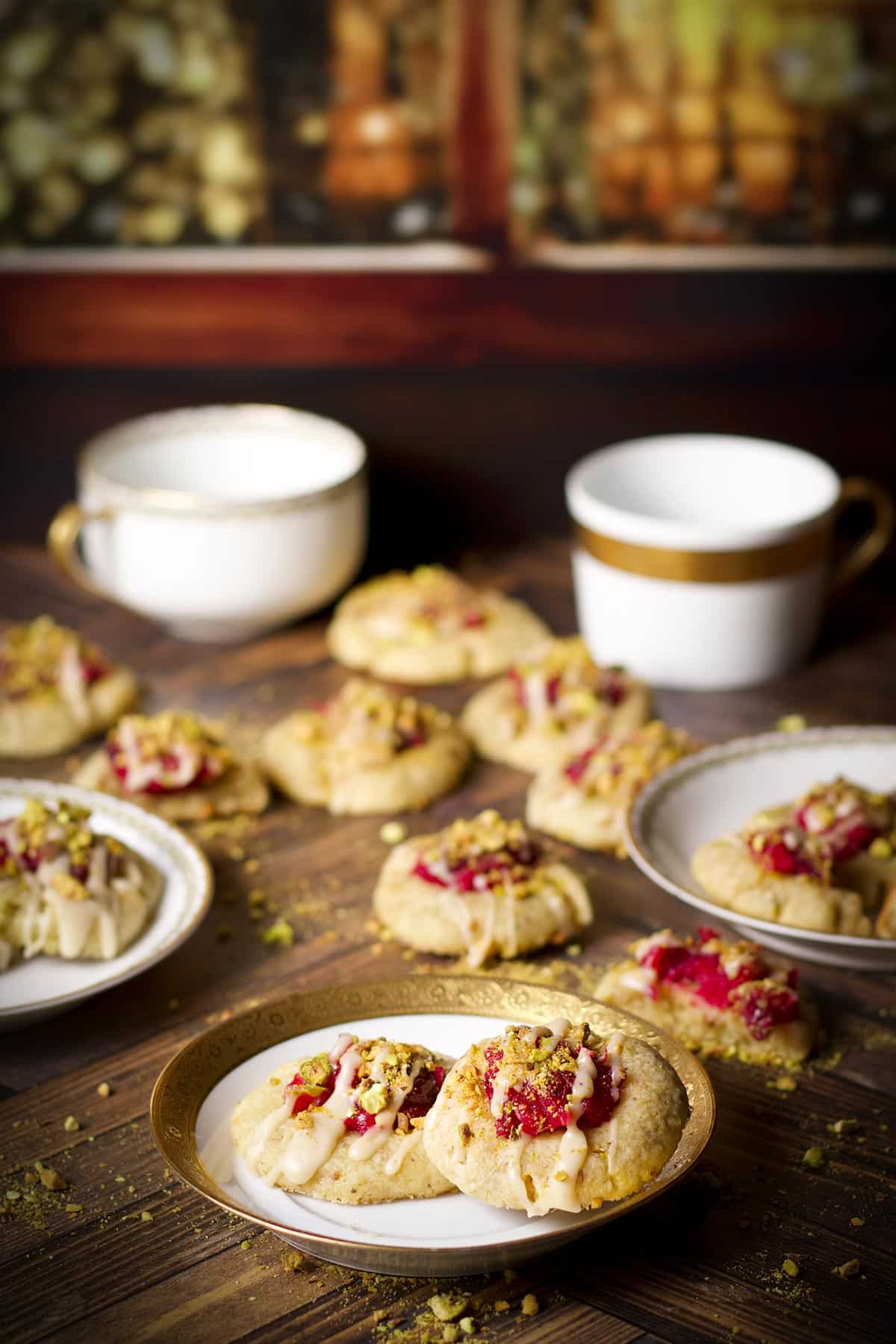 Several cranberry pistachio cookies scattered around a wooden table, some on plates and some just lying on the table.