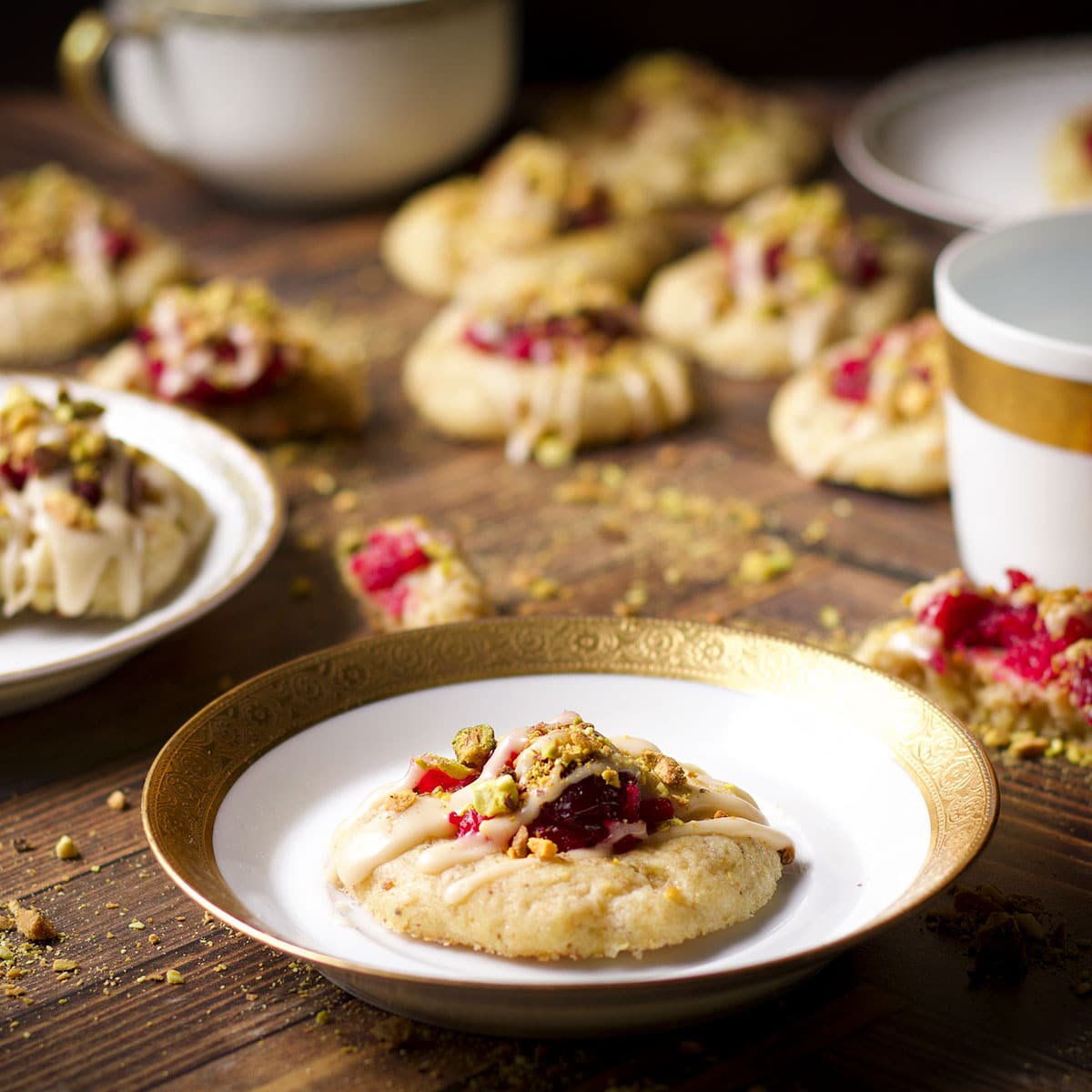 A cranberry pistachio cookie on a small gold-trimmed dessert plate with more cookies on the table all around the plate.