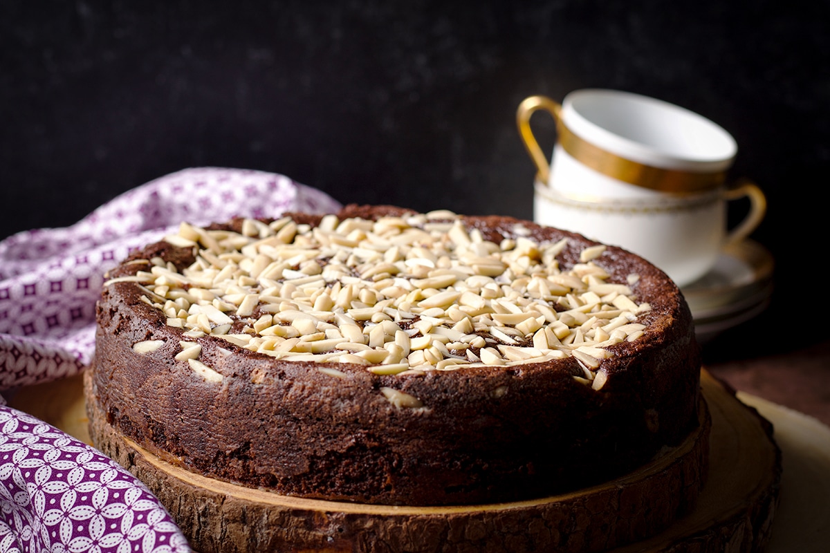 A freshly baked chocolate ricotta cake covered in almonds and resting on a wood cake board.