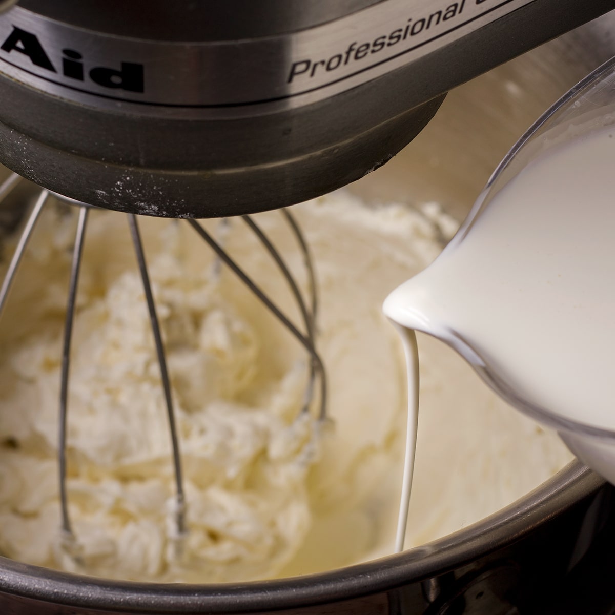 Pouring heavy cream into the bowl of an electric mixer while it beats mascarpone frosting.