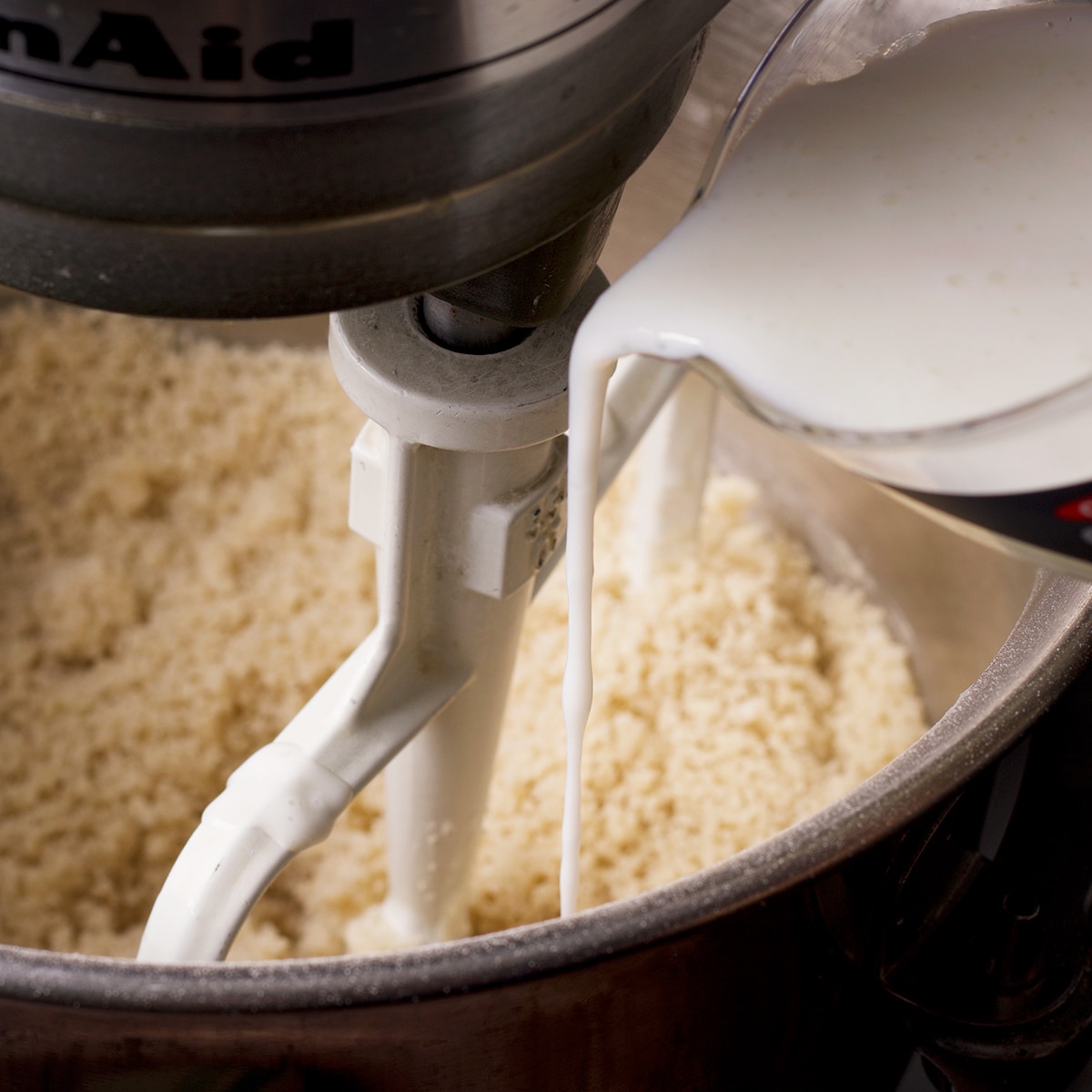 Pouring buttermilk into cake batter while an electric mixer is beating the batter.