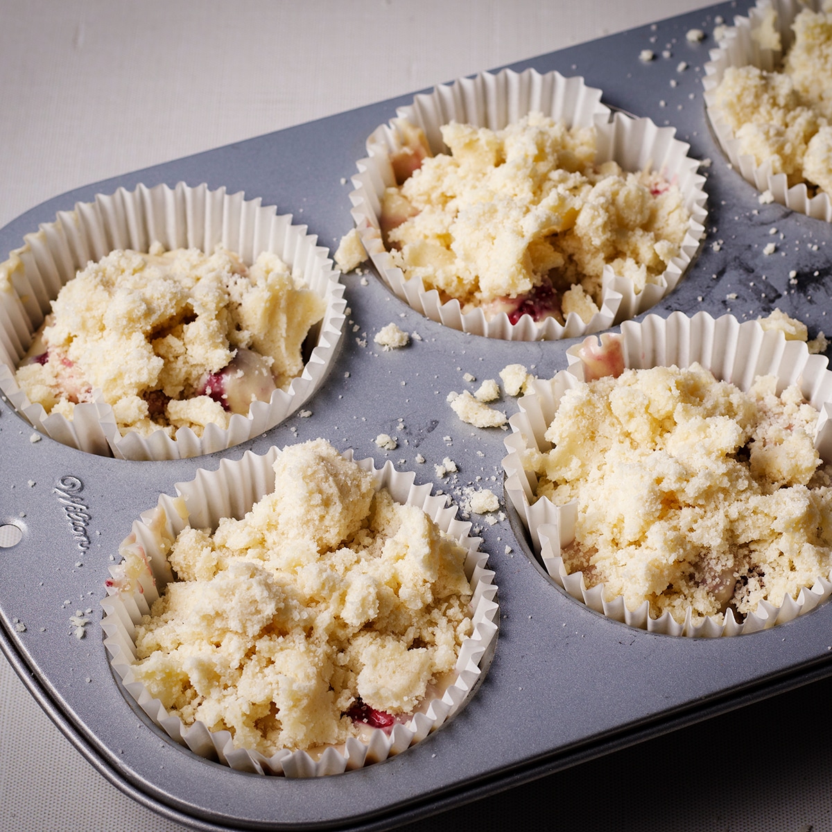 A jumbo size muffin tin fitted with paper liners that are filled with raspberry muffin batter topped with crumb topping.