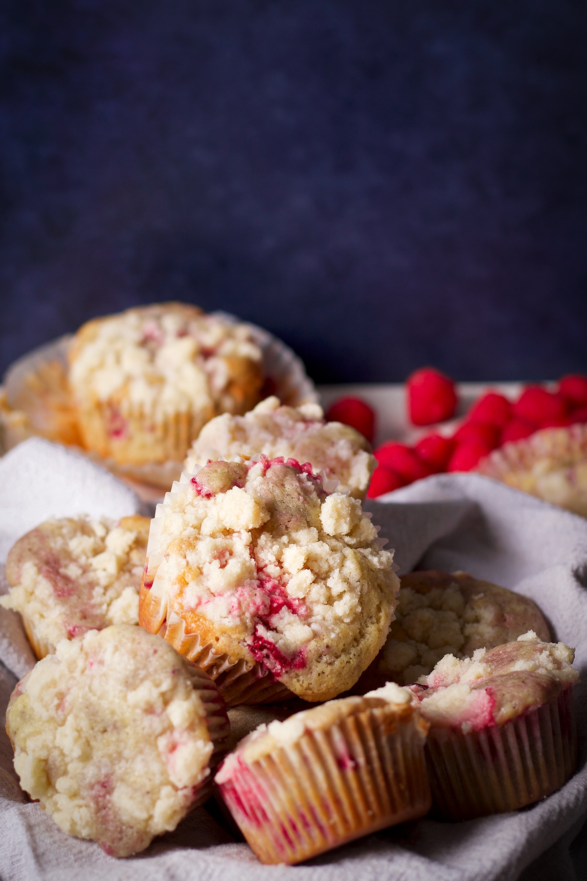 A basket of raspberry muffins on a white table with a pile of fresh raspberries behind the basket.