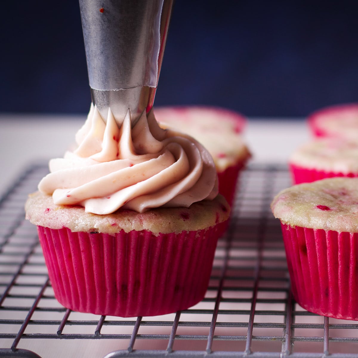 Using a pastry bag and cupcake tip to pipe a swirl of raspberry cream cheese frosting on a raspberry cupcake.