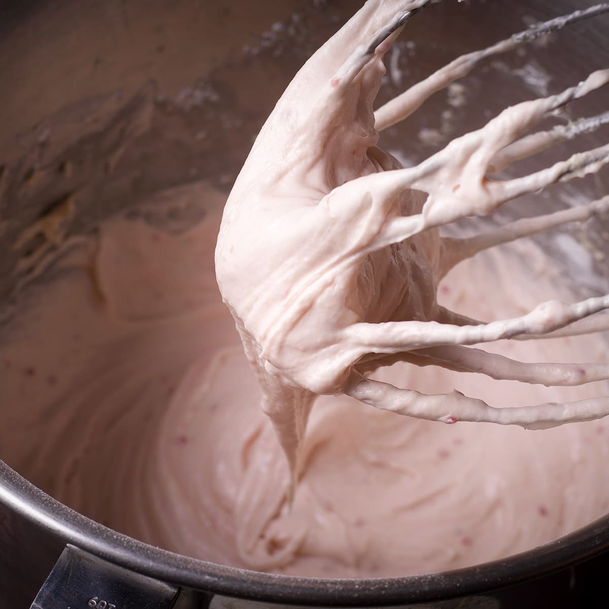 Raspberry cream cheese frosting dripping from the whisk attachment of an electric mixer into a mixing bowl.