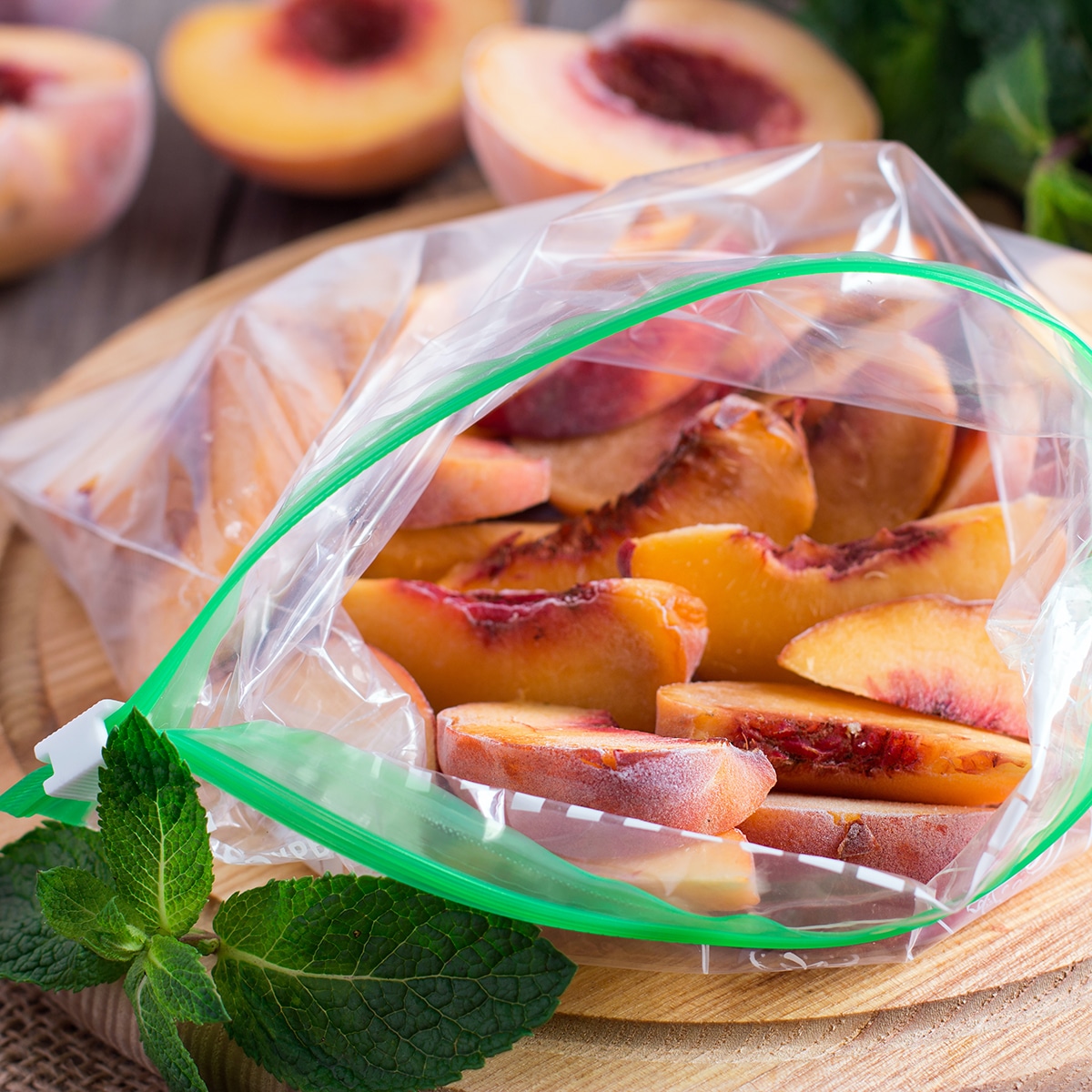 Frozen peach slices with the skin on in a zip-top bag on a table next to frozen peaches that have been cut in half.