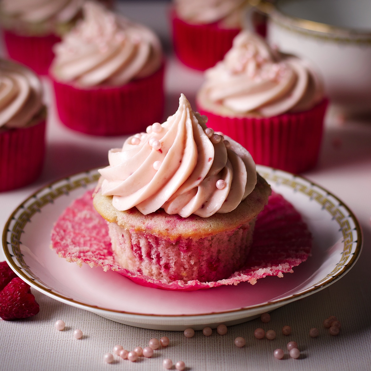 A raspberry cupcake on a dessert plate with several other cupcakes resting on the table behind it.