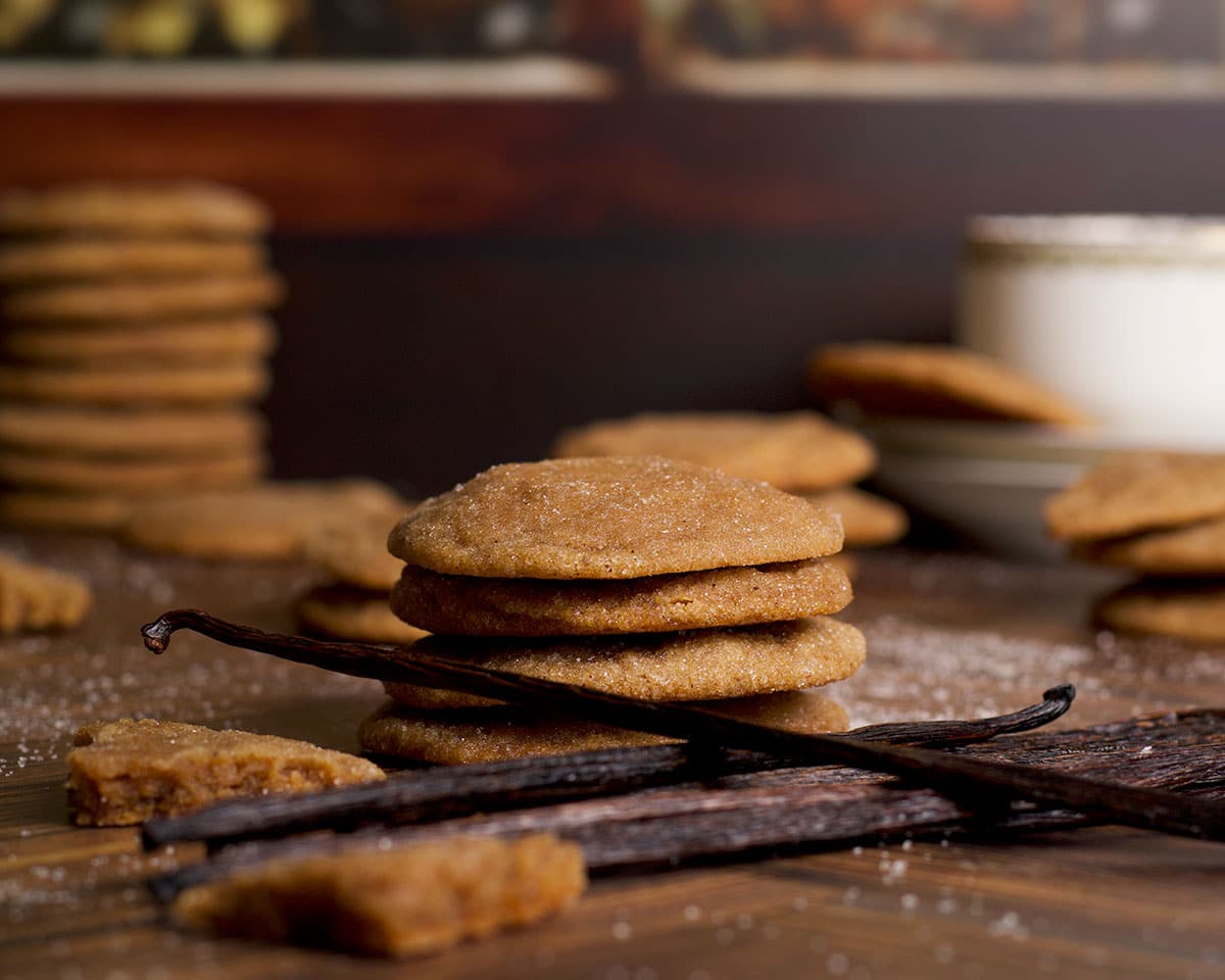 A pile of vanilla beans resting on a table next to stacks of brown sugar vanilla bean cookies.