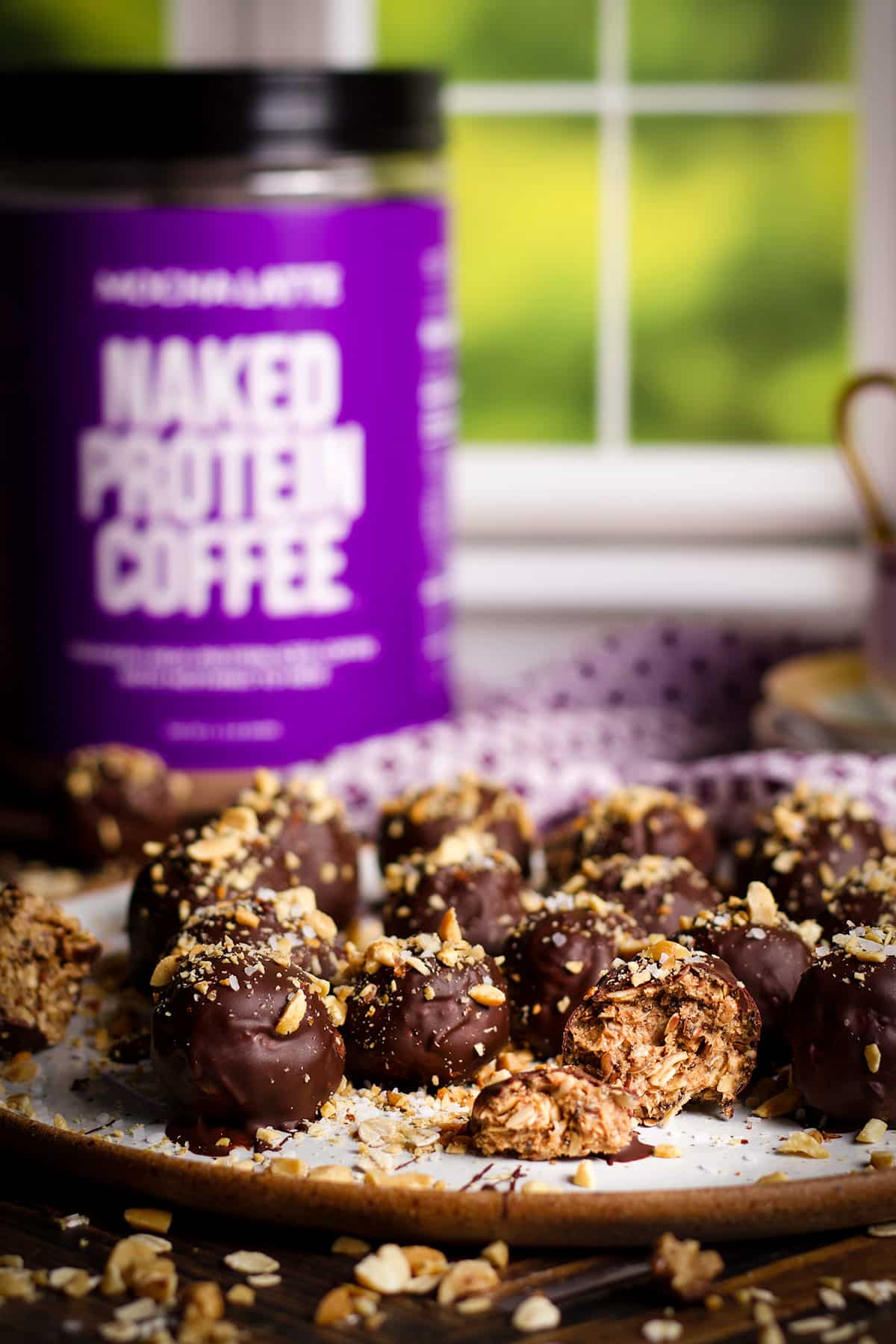A plate of chocolate covered peanut butter and oat protein balls next to coffee cups and a container of Naked Protein Coffee.